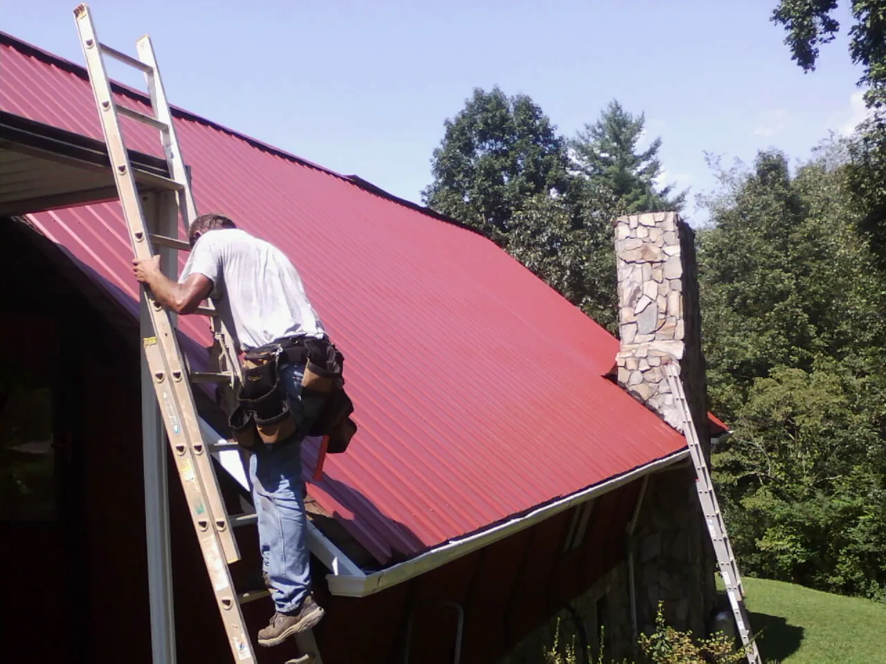 Roofer on a ladder beside a red metal roof and stone chimney — Soffit & Fascia Repair by Roofing Brothers in Arispe